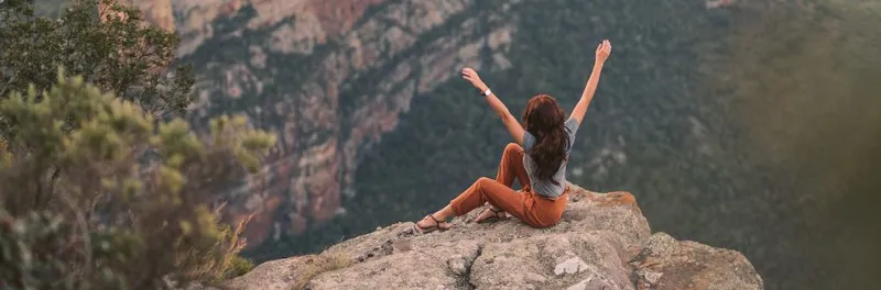 woman wearing a grey t-shirt and brown trousers sat on mountain top over looking the lake